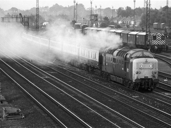 Black-and-white elevated view of Class 55 Deltic 55018 Ballymoss accelerating past Dringhouses yard, York, with a passenger train, its exhaust drifting across the main running lines and vans standing in the yard beyond