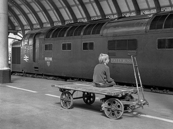 Black-and-white photograph of Class 55 Deltic 55018 Ballymoss standing beneath York station canopy, watched by a lone trainspotter seated on a platform trolley