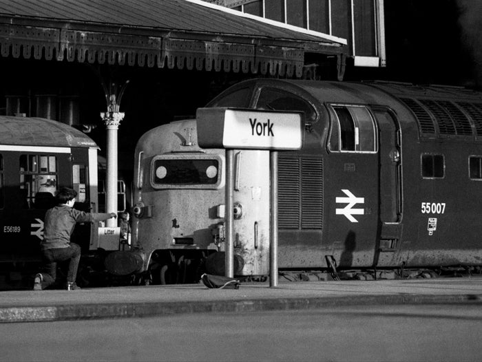 Black-and-white photograph of a young trainspotter kneeling on the platform at York, arm outstretched towards Class 55 Deltic 55007 Pinza beneath the ornate station canopy, with the York nameboard and his bag in the foreground