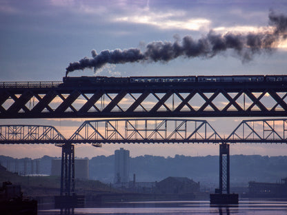 Colour telephoto photograph of Flying Scotsman hauling the Rail 150 Steam Special across the King Edward Bridge at sunset, trailing dark smoke above the River Tyne