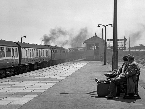 Black-and-white photograph of Class 52 D1047 Western Lord pulling away from Par station beneath a plume of exhaust, with two passengers seated on a platform bench